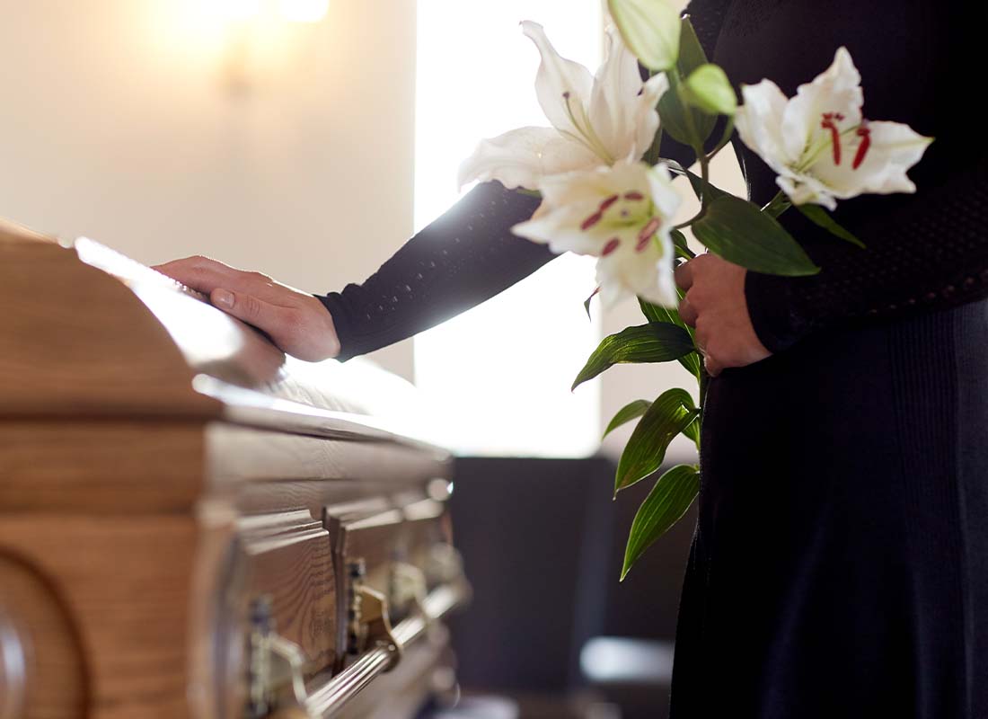 Funeral Home Insurance - Close-up of Woman Holding Lily Flowers While Other Hand Is Resting on a Coffin at a Funeral in a Funeral Home